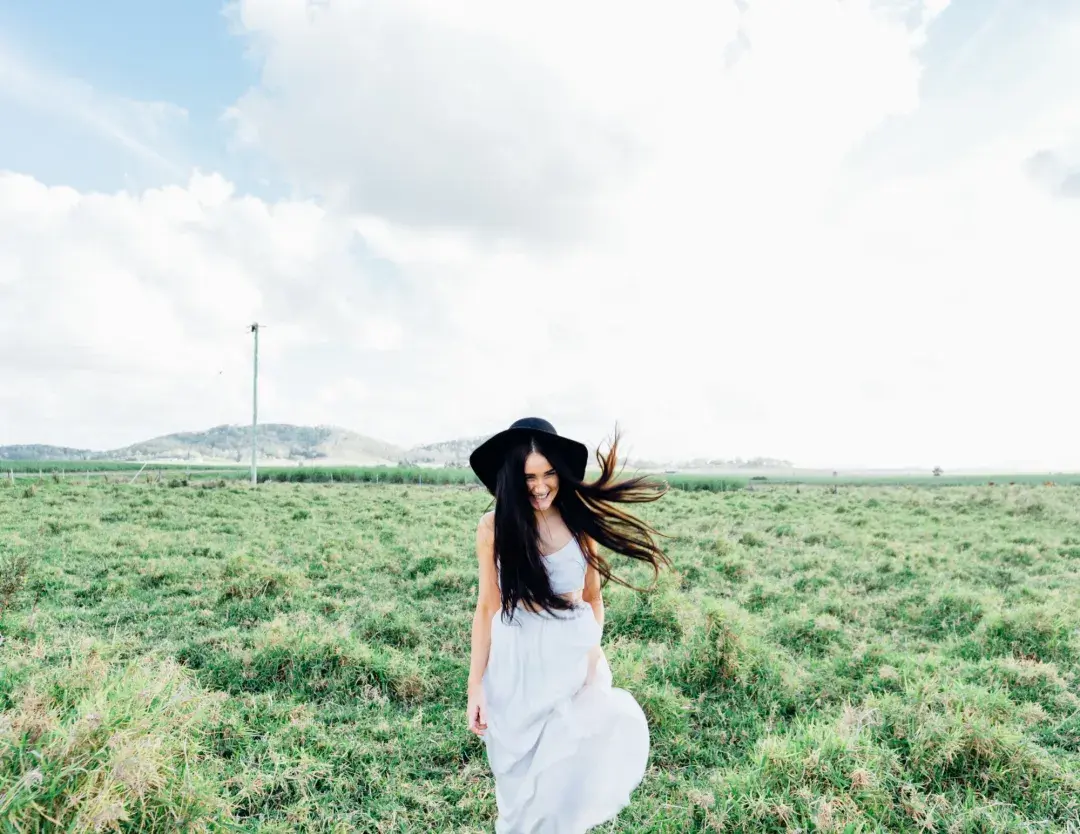 Woman with white dress, long dark hair, a black hat walking towards the viewer with a grass field background