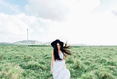 Woman with white dress, long dark hair, a black hat walking towards the viewer with a grass field background